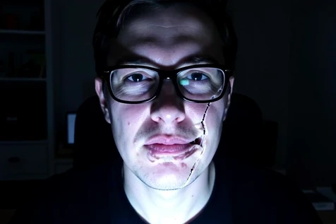 Close-up of a person wearing thick black-framed glasses on a webcam during a face swap that is visibly failing, the swapped face smearing and tearing around the mouth and breaking at the glasses frame edge, leaving a doubled, glitchy seam across the cheek. The subject sits in a dim home-office at night. Single harsh white monitor glow lights the face from below and front, cool and uneven, deepening shadows under the eyes and exaggerating the broken blend. Glitchy, unflattering, diagnostic mood.
